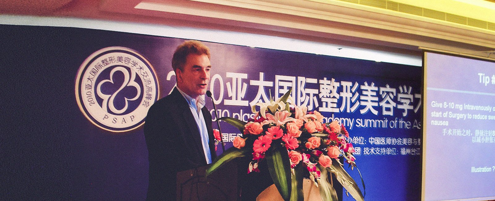 A suited man speaks at a conference podium with a "PSAP" banner and floral decoration.