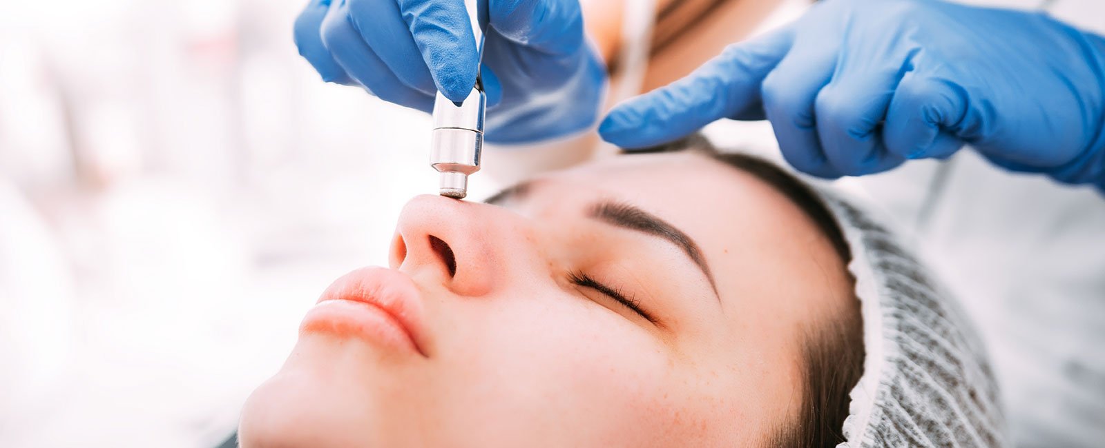 plastic-surgery-hawaii-hero-1 A professional performs dermabrasion on a person's nose in a clinical setting; person wears a hair cover.