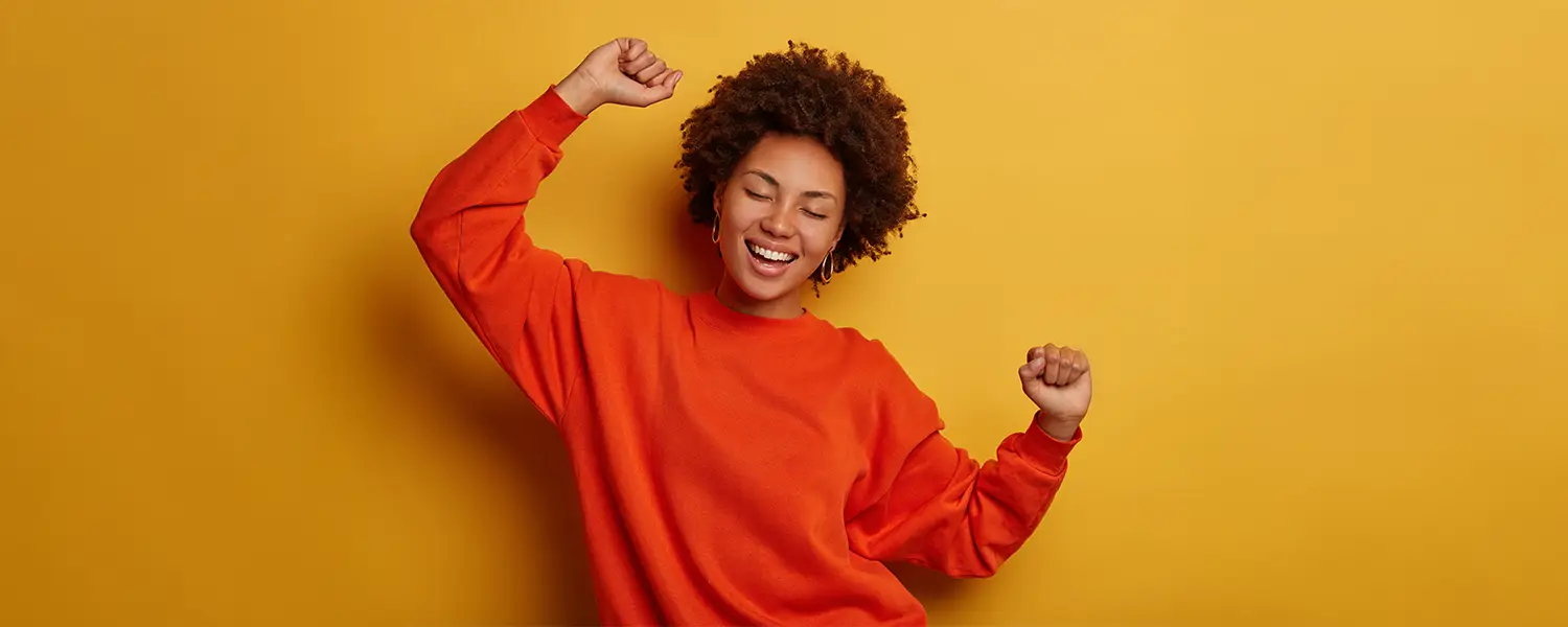 Curly-haired person in orange sweater smiles with eyes closed