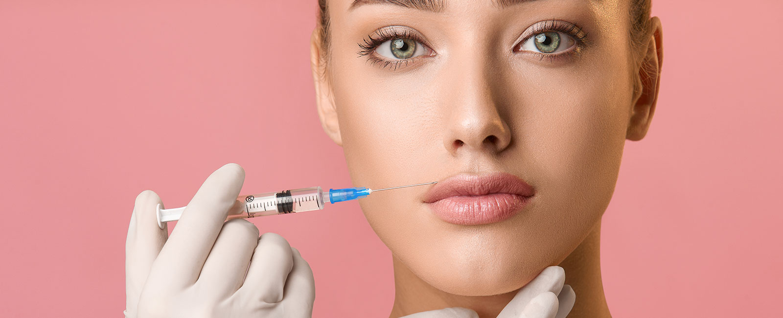Person with light skin and green eyes receives lip augmentation; syringe held near lips against pink background.