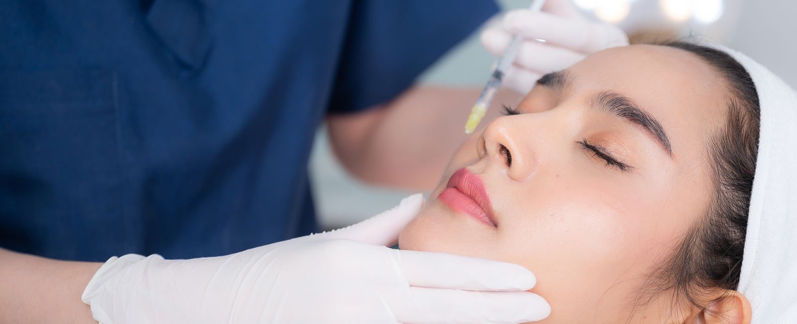 plastic-surgery-hawaii-hero-1 A healthcare professional in scrubs injects a patient's cheek in a clinical setting.
