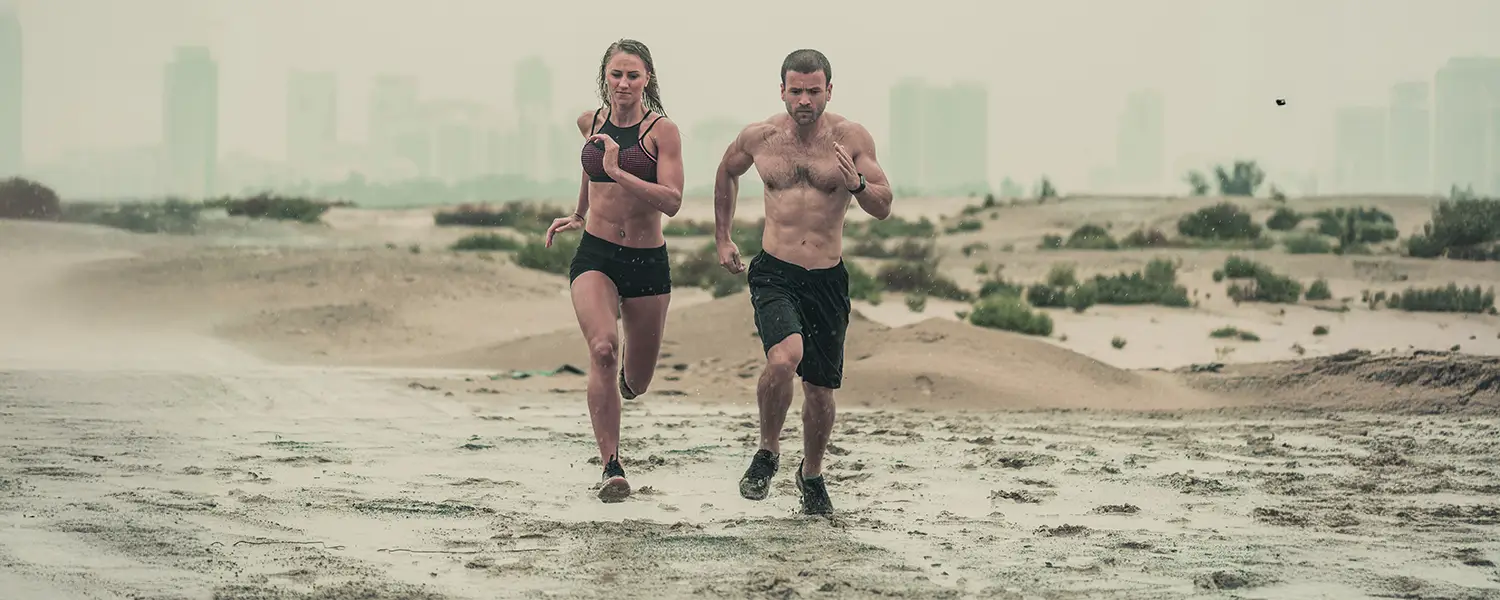 Two people run on a wet beach with a city skyline; overcast sky