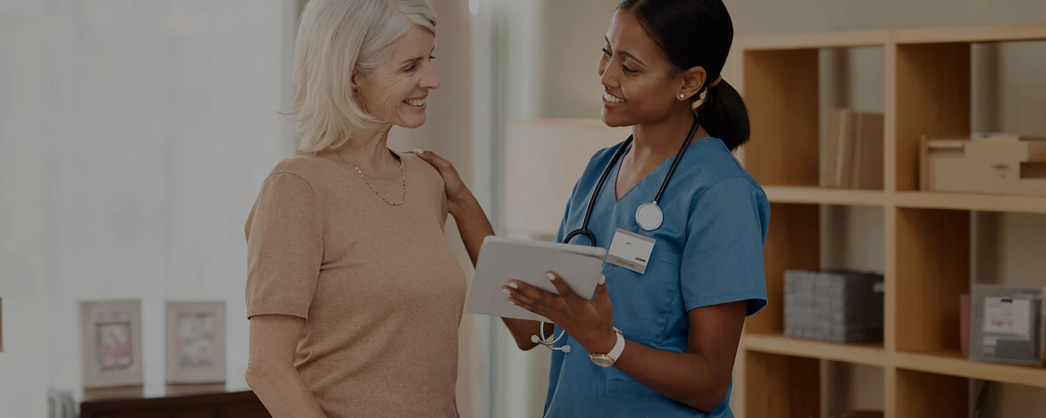 A healthcare worker in blue scrubs talks to an older woman in a well-lit room.
